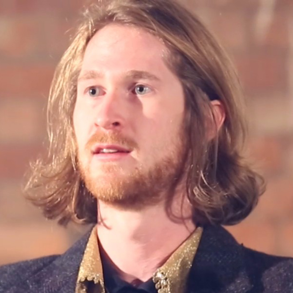 A photo of Andy Owen Cook, a white man with shoulder length blonde hair and a beard. He wears a yellow shirt with a grey jacket, and looks off to the left of the frame.