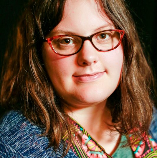 A photo of Sarah Gonnet, a white woman with long brown hair, wearing red classes with red frames as well as a green and blue top and smiling at the camera.