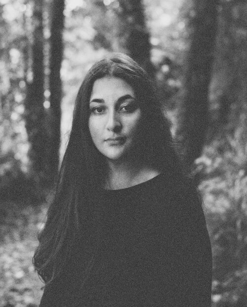 A black and white photo of Lydia Hounat, a British-Algerian woman with long dark hair, wearing a black top. She is standing in some woodland, and looking straight at the camera.