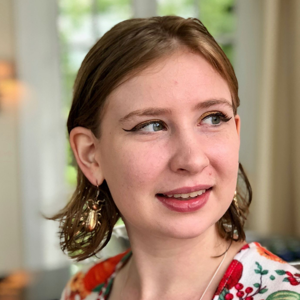 A photo of Sarah Fletcher, a white woman with mid-length light brown hair pushed back behind her ears, wearing a floral top. She is smiling and looking off to the right of the frame.