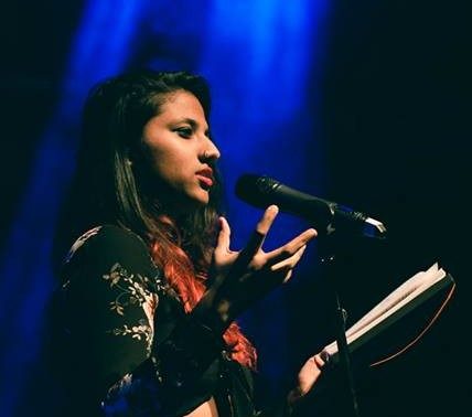 A photo of Prerana Kumar, an Indian woman with long brown hair swept to one side, wearing a dark patterned top, holding a notebook and speaking into a microphone, with a blue light behind her.