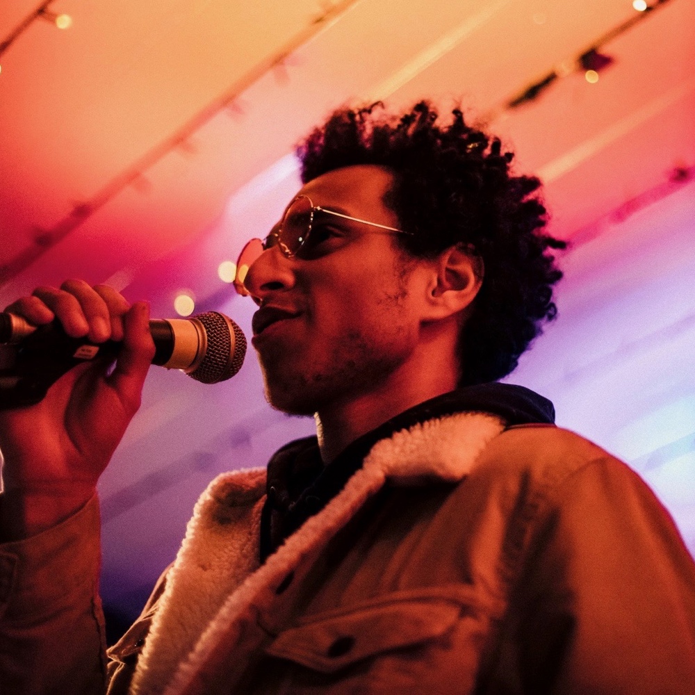 A photograph of Fahad Al-Amoudi, a man of Ethiopian and Yemeni heritage with a short beard and curly black hair. He wears glasses and a brown jacket with a sheepskin lining, and is speaking into a microphone.