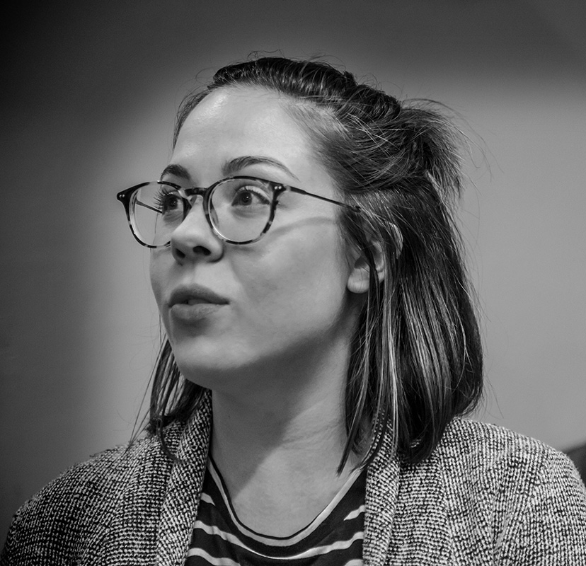 A greyscale photo of Laura Attridge, a white woman with shoulder length brown hair. She wears her hair half-up-half-down, and is wearing glasses and a knitted cardigan over a striped t-shirt. She looks off to the left of the frame.