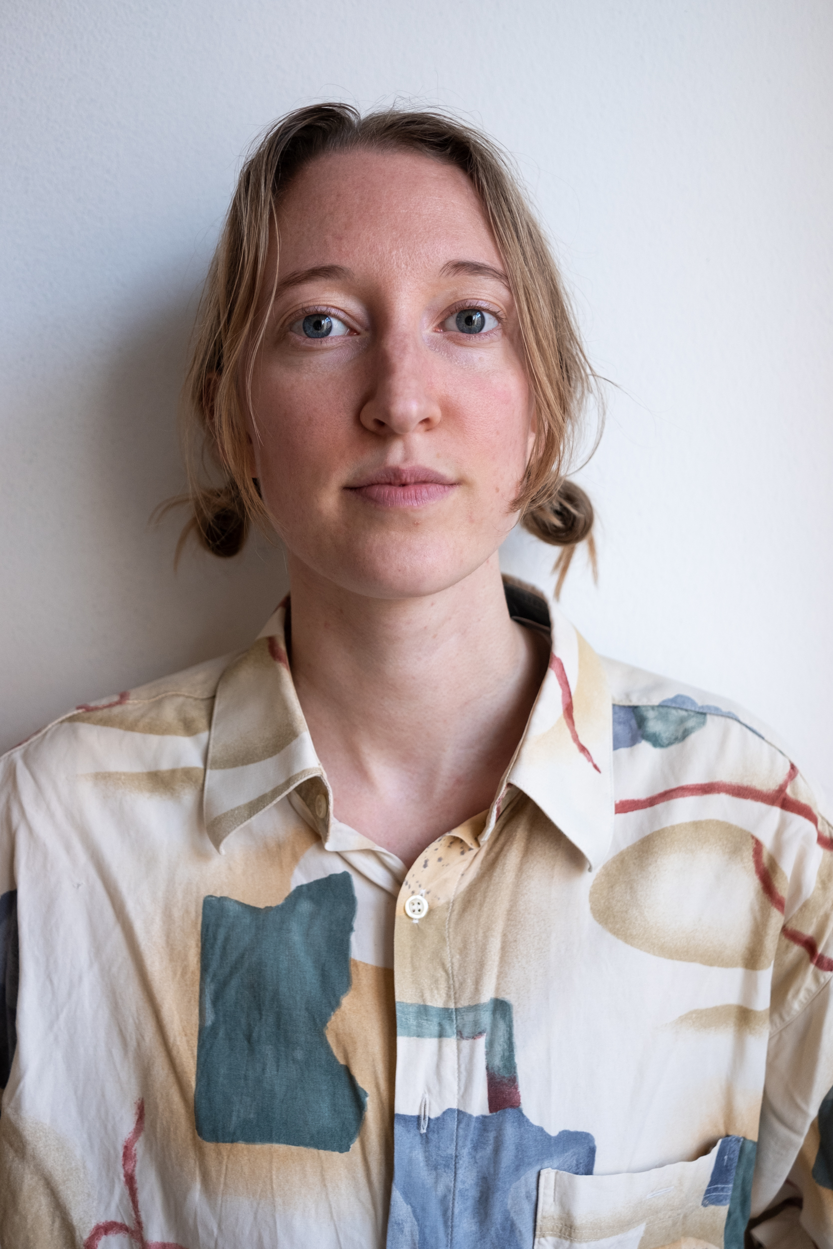 A photo of Louise Essex, a white woman with mind-length blonde-brown hair tied back in bunches. She stairs straight at the camera, and is wearing a pale cream coloured shirt with abstract blue shapes on it.