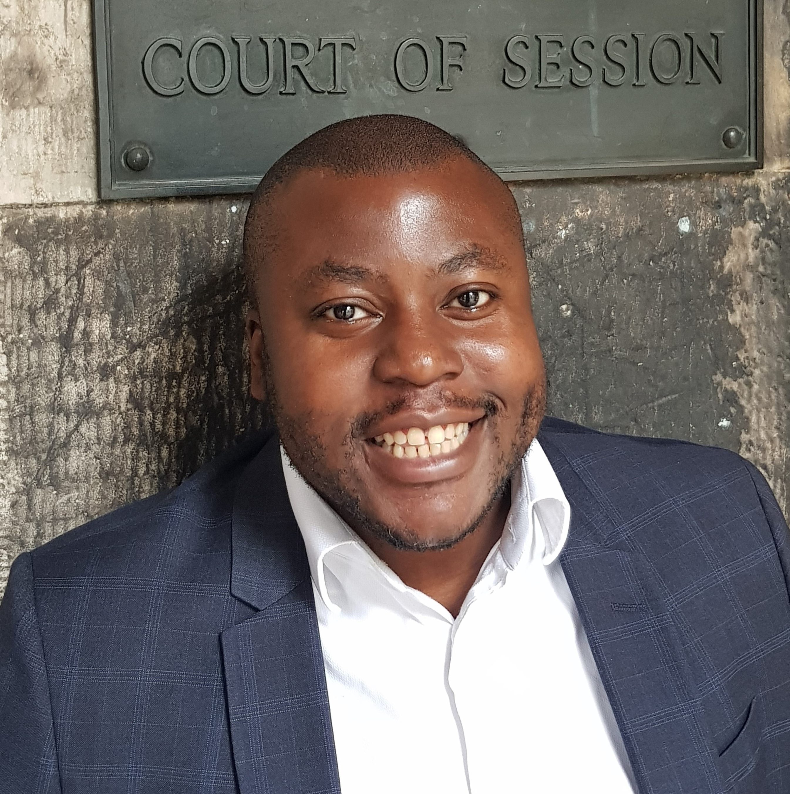 A photo of May Sumbwanyambe, a black man wearing a white shirt and grey blazer. He smiles at the camera, and is stood in front of a stone wall. A sign on the wall reads 