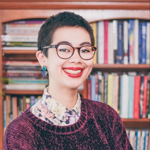 A photo of Helen Bowell, a half-Chinese woman with short dark hair, wearing glasses, a floral shirt and purple jumper. She smiles at the camera, standing in front of a bookcase.