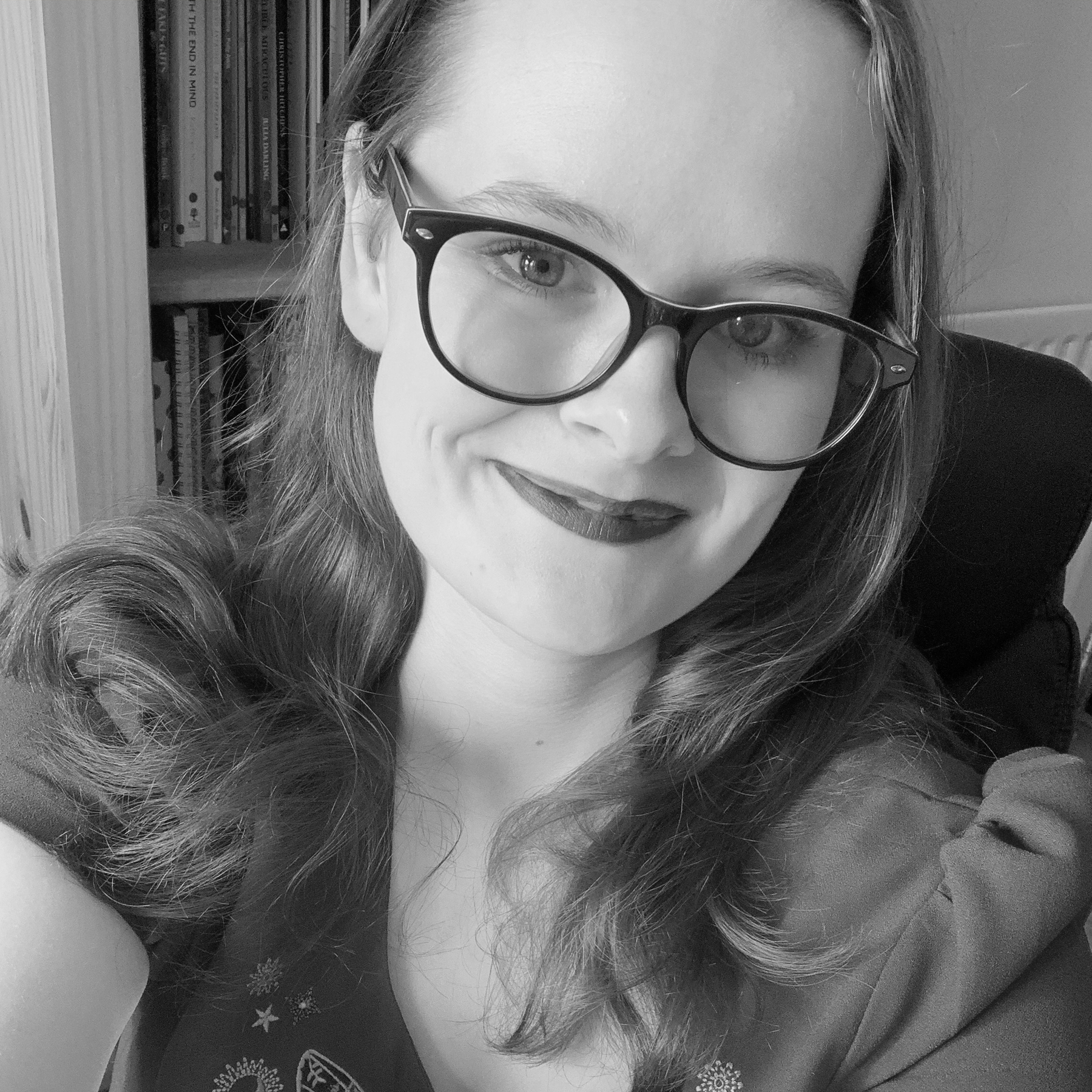 A black and white photo of Hannah Hodgson, a white woman with long light brown hair and glasses. She is smiling at the camera, and wearing dark lipstick.