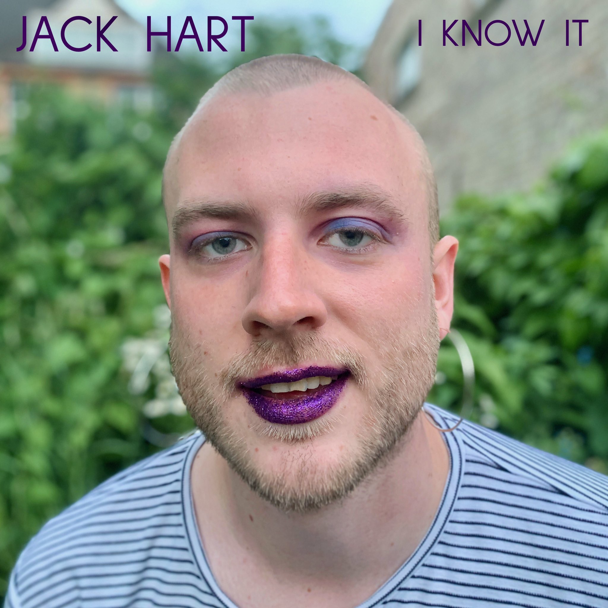 A photo of Jack Hart, a white non-binary person with closely shaven hair and a blonde beard. They are wearing a purple lipstick and a stripey t-shirt, and are smiling at the camera.