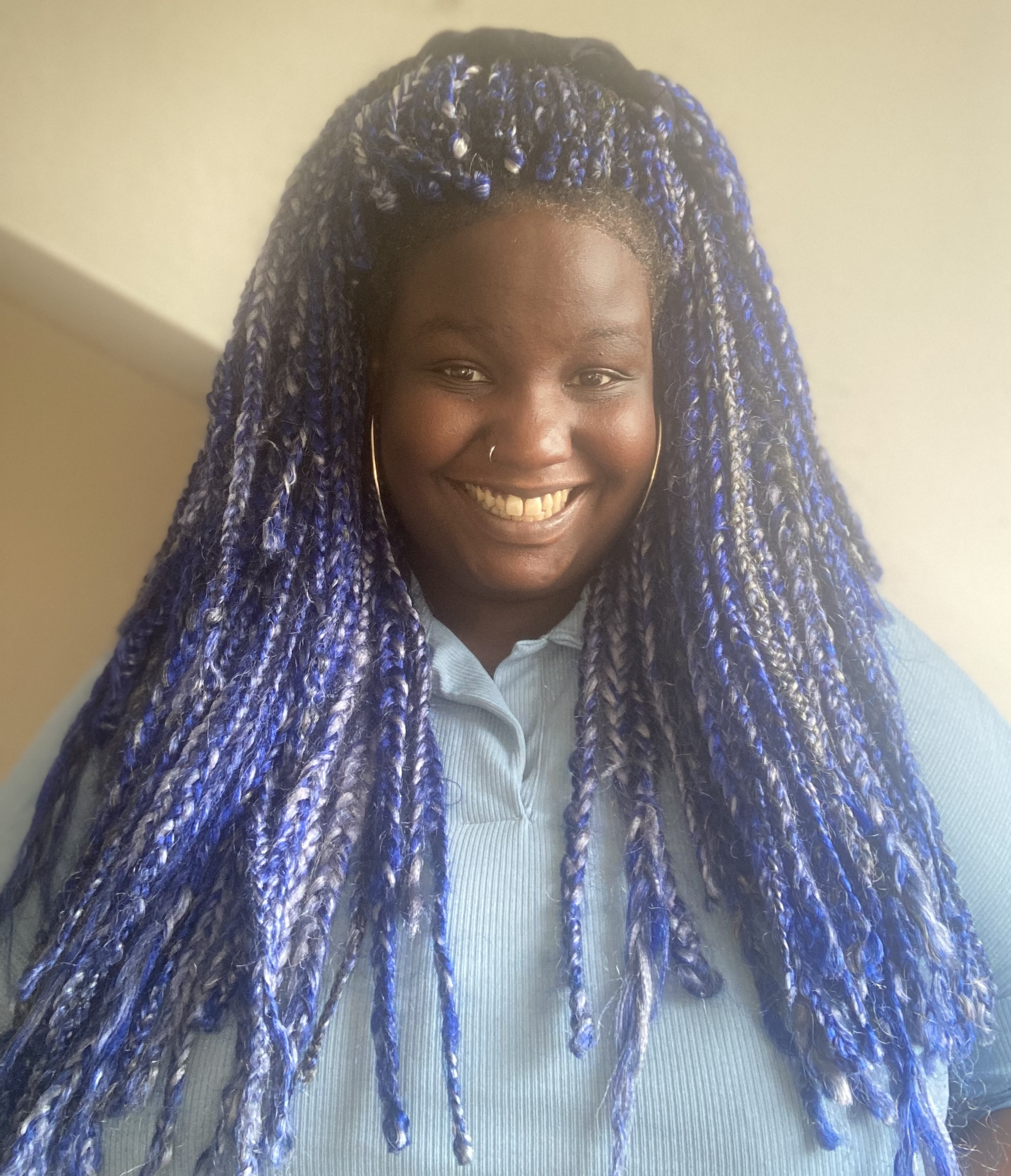 A photo of Ruth Awolola, a black woman with long blue, purple and grey braids and wearing a light blue shirt. She smiles at the camera.