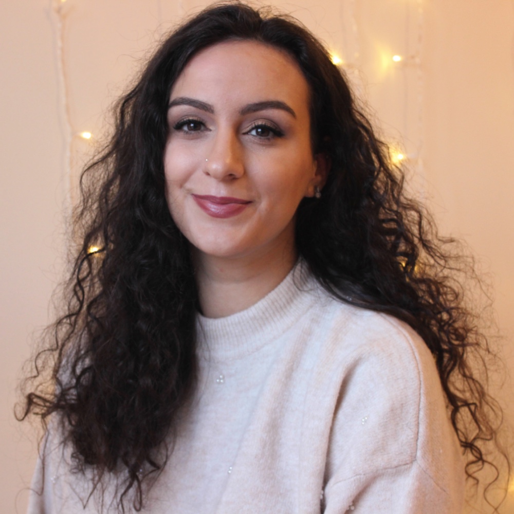 A photo of Nasim Asl, a British-Persian woman with long, dark wavy hair. She wears a cream jumper and smiles at the camera. Behind her, strings of fairylights are hanging on the wall.