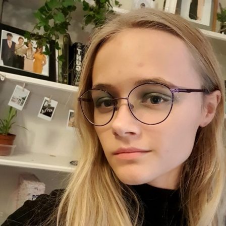 A photograph of Lauren Hollingsworth Smith, a white woman with long blonde hair. She wears glasses and a black top, and stands in front of a bookshelf.