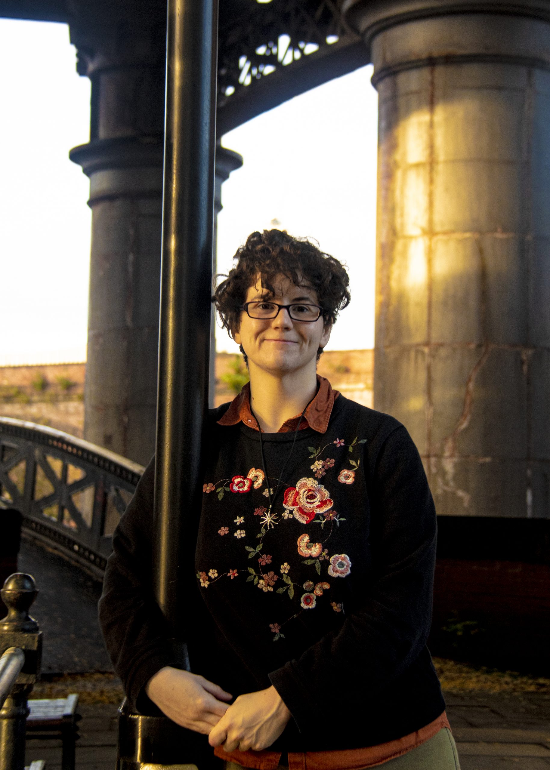 A photograph of Elizabeth Gibson, a white woman with brown curly hair. She is wearing glasses and a black jumper with flowers on it. She is leaning against a lamppost.