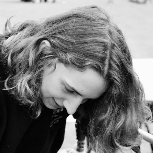 A black and white photo of Alice Noel, a white woman with shoulder length wavy hair, wearing a black top and leaning to look down the right hand side of the frame.