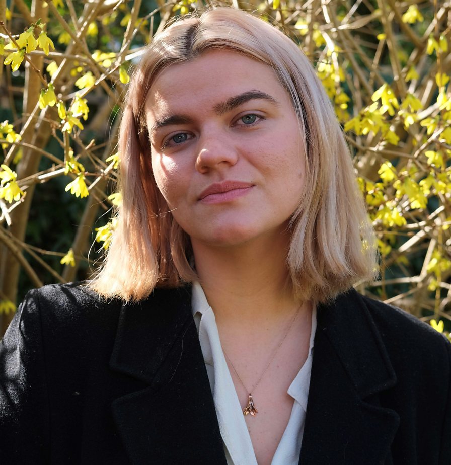 A photo of Saba Sams, a white woman with shoulder length blonde hair, wearing a white shirt under a black jacket, and stood outside.