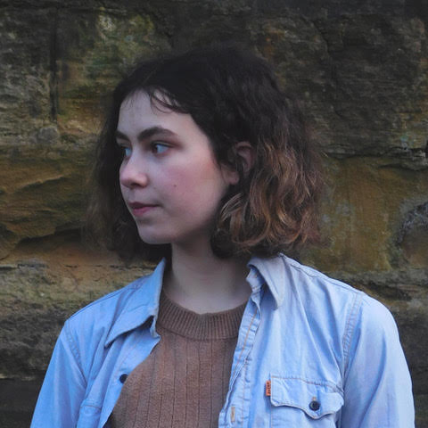 Liv Aldridge, a white woman with short dark hair in a denim shirt, looks to the left hand side of the frame on a nature background.