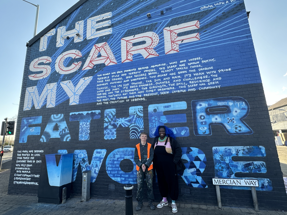 Graffiti artist Oskar with a K and poet Ruth Awolola stand in front of the gable end of a two story building emblazoned with the Stockport County motto The Scarf My Father Wore, in blue of course, and the text of a collaborative poem.
