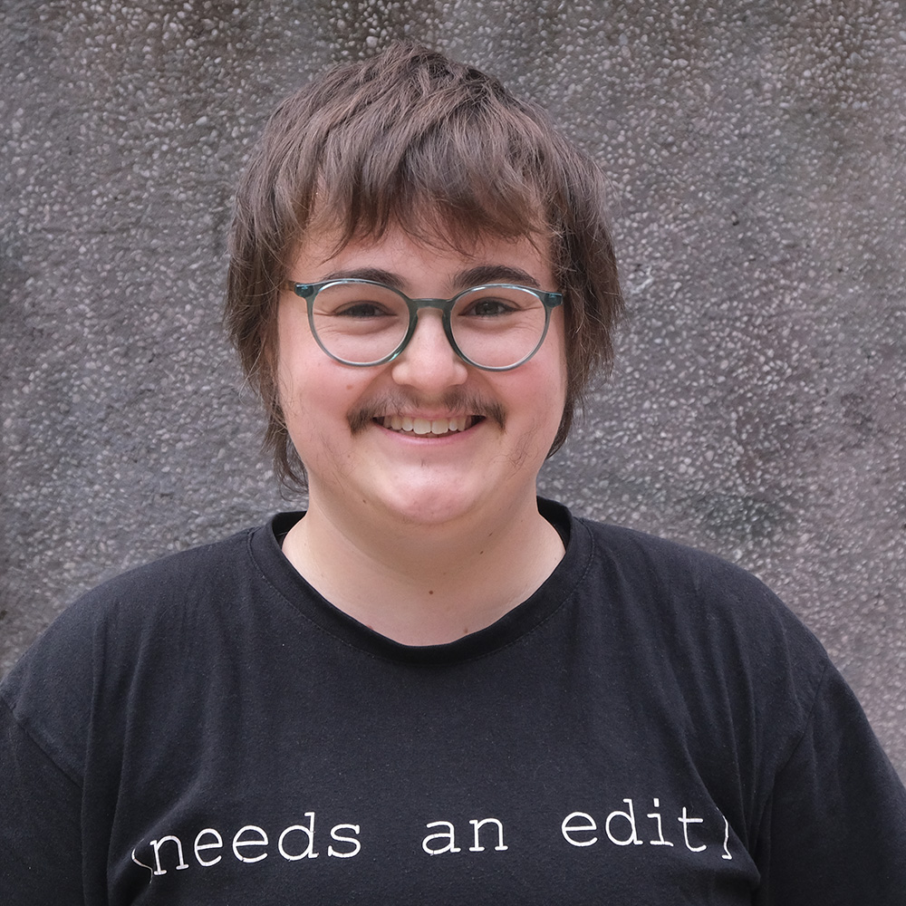 Headshot of Roma smiling and wearing a black sweat shirt that says Needs an Edit